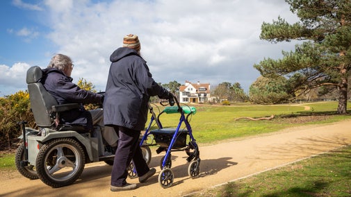 A visitor in a tramper next to a companion with a mobility aid walking towards Tranmer House at Sutton Hoo